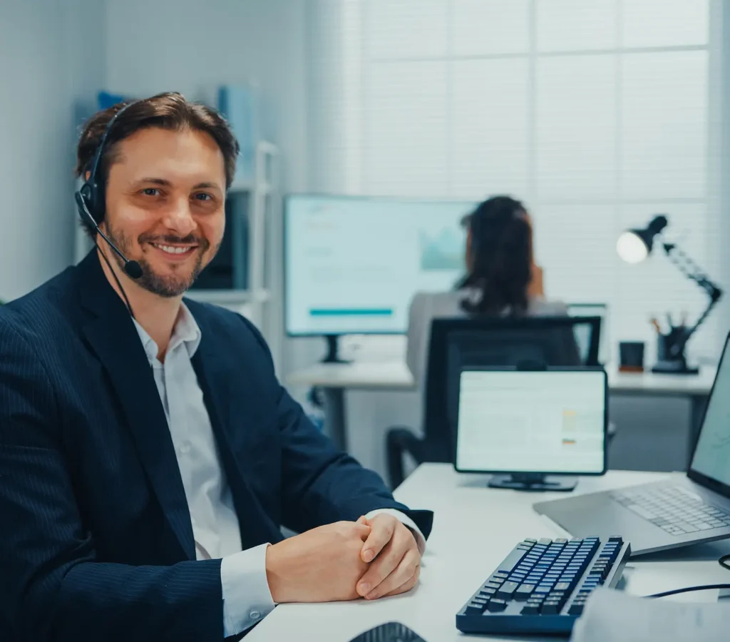 Smiling male call center agent wearing a headset looks at the camera while working at desk in a modern office. Phone consultation, client assistance, customer support and contact center service.