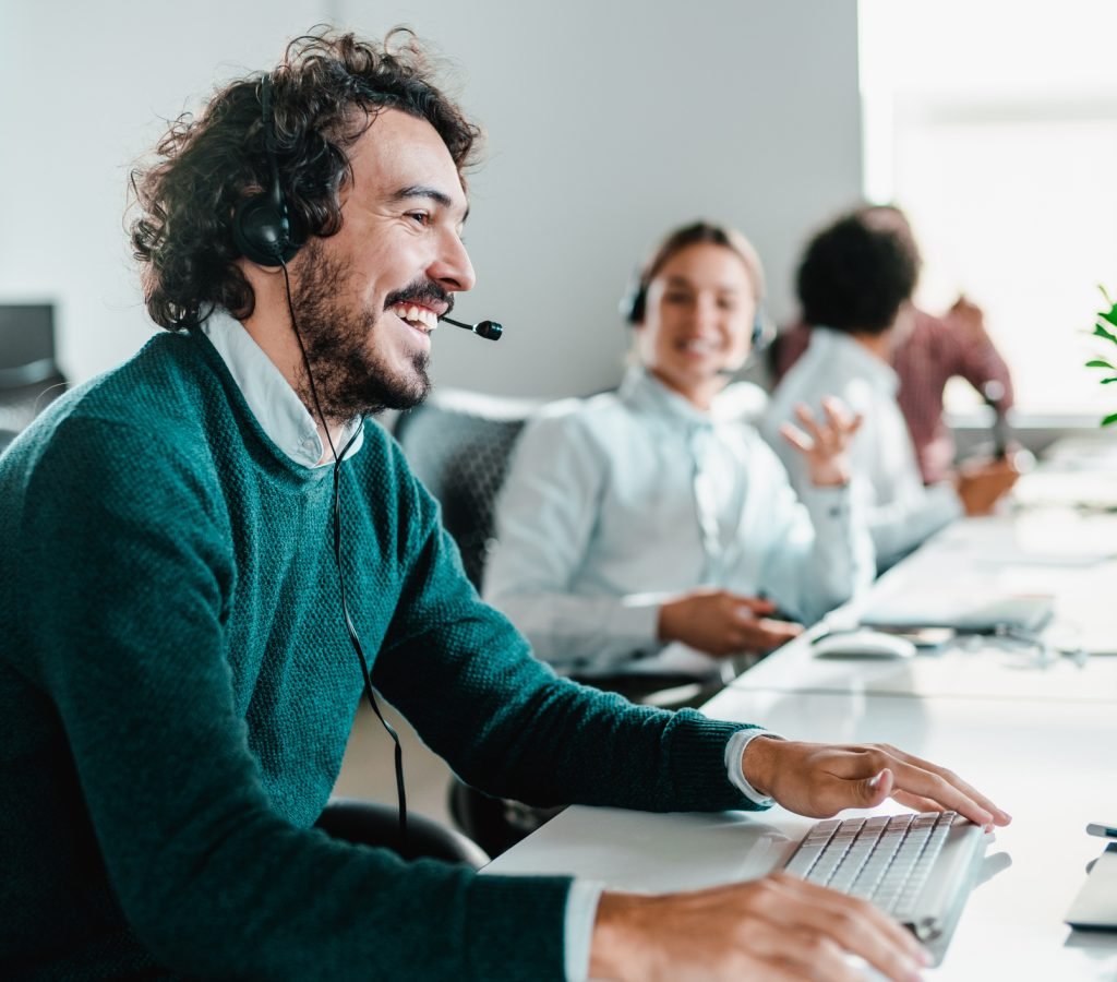 Smiling handsome young businessman working in call center. Shot of a cheerful young man working in a call center with his team. Confident male operator is working with colleagues. Call center operators sitting in a row at desks.